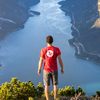 Wanderer bekleidet mit rotem T-Shirt mit Logoaufdruck blickt auf einen See mit Bergen ringsum